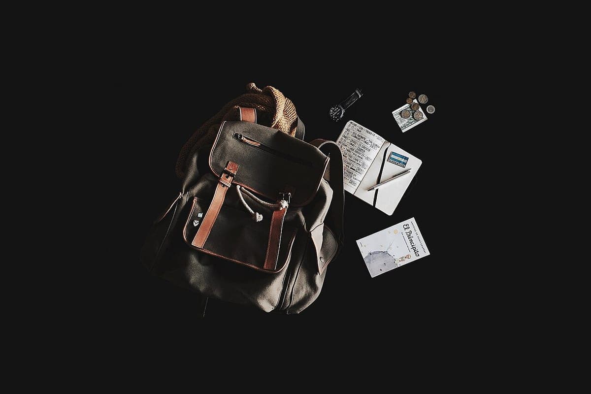 A canvas travel backpack laid out with a notebook, a watch and loose change